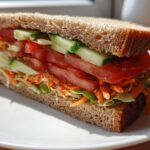 A tall Ultimate Veggie Sandwich made with whole wheat bread, stacked high with lettuce, shredded carrots, tomato slices, and cucumber.