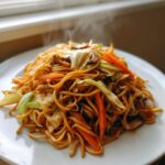 A close-up of steaming hot Vegetarian Chow Mein noodles mixed with bright orange carrots, green cabbage, and mushrooms on a white plate.