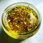 Overhead close-up of a bright yellow liquid containing chili flakes, garlic pieces, and herbs, representing the Chili Lime Marinade.