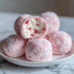 A stack of pink, powdered Watermelon Mochi balls, one cut open revealing a creamy white filling with red watermelon pieces.