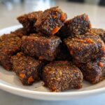 A pile of dark brown, textured, square-shaped homemade Werewolf Kibble treats stacked on a white dish.