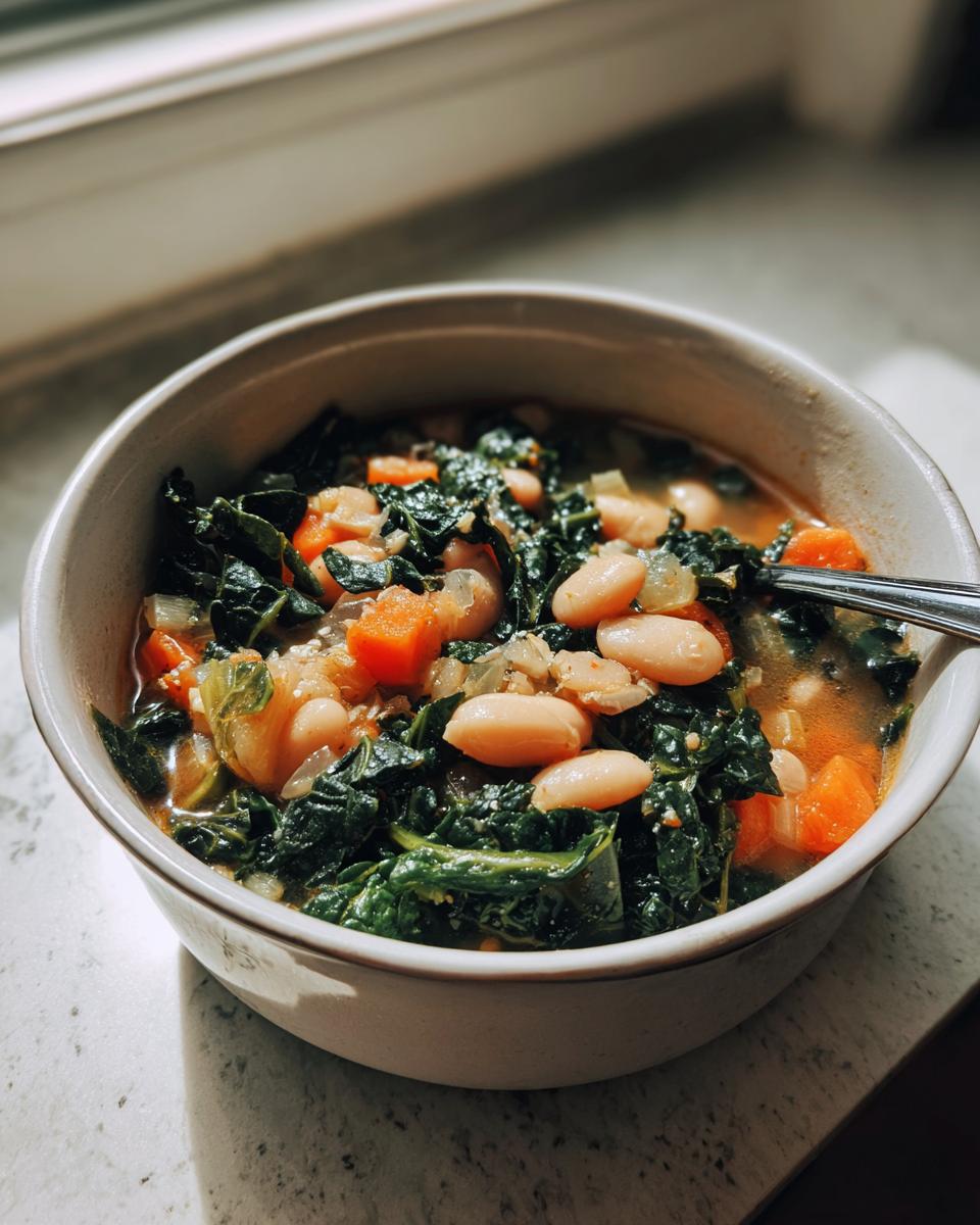 A close-up of a bowl filled with White Bean Kale Soup, showing white beans, dark green kale, and diced carrots in broth.