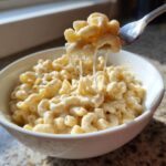 A fork lifts macaroni from a bowl of creamy White Cheddar Mac And Cheese, showing long, satisfying cheese pulls.