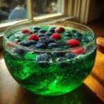 Close-up of bright green, bubbly Witch Potion Punch garnished with floating blueberries and raspberries in a clear glass bowl.
