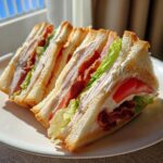 Three quarters of a classic Club Sandwich stacked on a white plate, showing layers of turkey, bacon, lettuce, tomato, and mayonnaise.