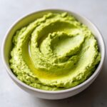 Close-up of creamy, bright green Edamame Hummus swirled in a white bowl, drizzled with olive oil.