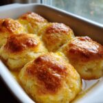 Close-up of freshly baked Butter Swim Biscuits golden brown and glistening in a pool of melted butter in a white baking dish.
