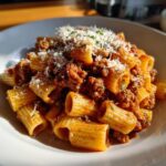 A close-up, sunlit serving of Italian Sausage Rigatoni pasta coated in a rich meat sauce and topped with grated Parmesan cheese.