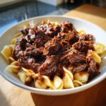A close-up of a white bowl filled with tender, shredded Short Rib Ragu served over shell pasta.