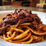 A close-up of a hearty serving of Short Rib Ragu piled high on thick, tubular pasta in a white bowl.