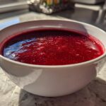 Close-up of a white bowl filled with deep red Spiced Cranberry Soup, glistening in natural light.
