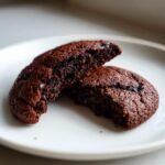 Close-up of a rich, dark chocolate Vegan Black Bean Cookie broken in half, showing its fudgy interior on a white plate.