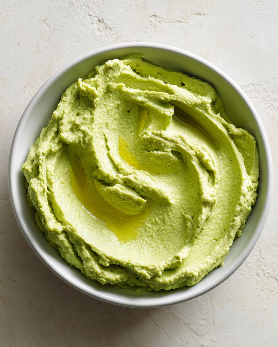 Overhead shot of creamy, bright green Edamame Hummus swirled in a white bowl, drizzled with olive oil.