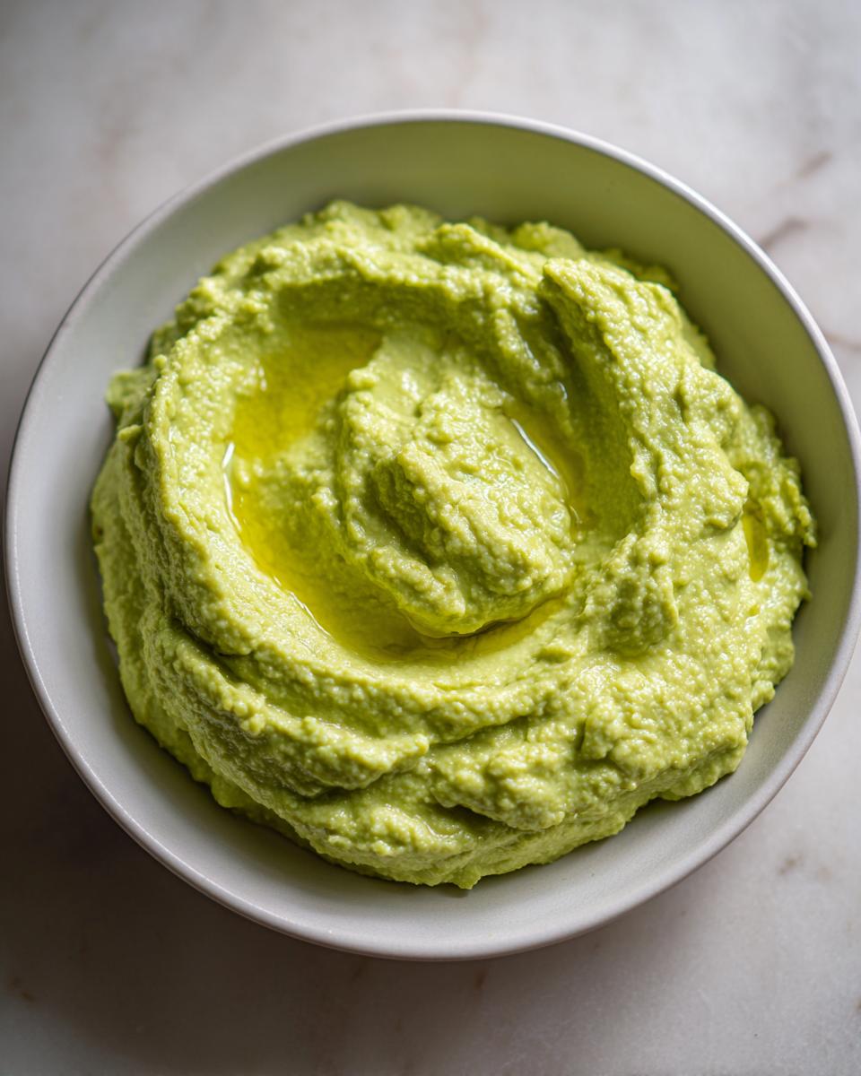 A close-up overhead shot of creamy, bright green Edamame Hummus drizzled with olive oil in a light gray bowl.